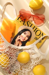 Photo of String bag with fresh lemons, fashion magazine and beach accessories on beige background, flat lay