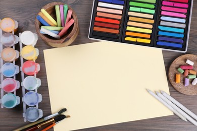 Blank sheet of paper, colorful chalk pastels and other drawing tools on wooden table, flat lay. Modern artist's workplace