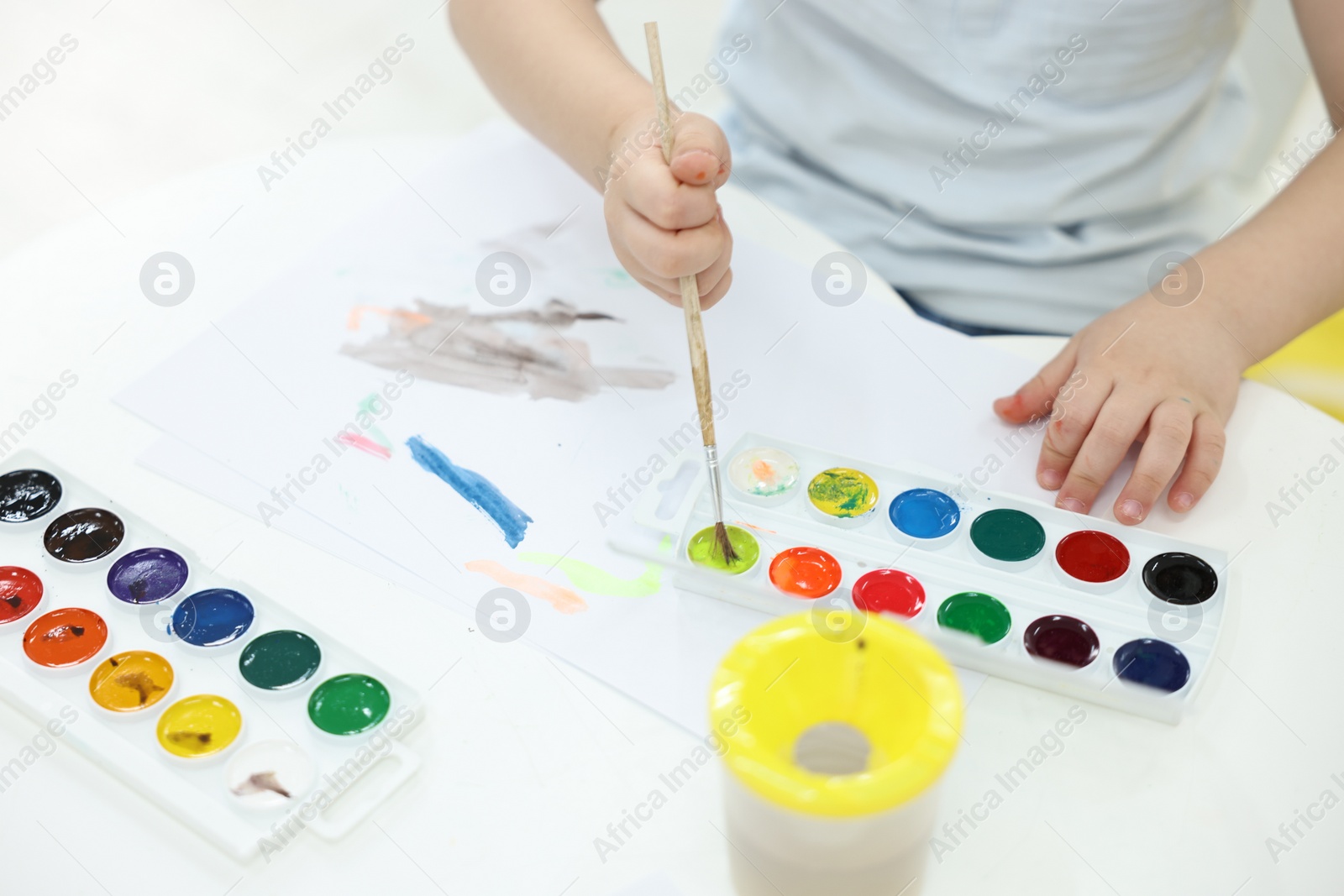 Photo of Little boy painting with brush and watercolor at white table, closeup