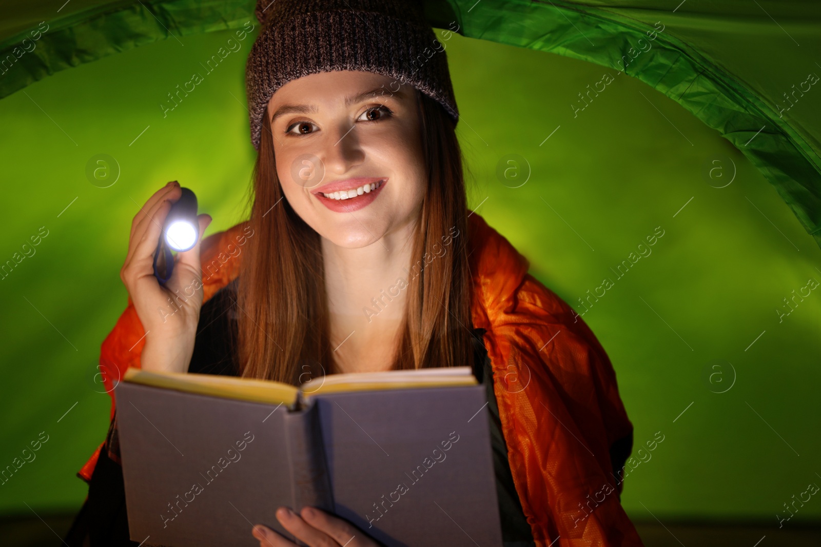 Photo of Young woman with flashlight reading book in tent