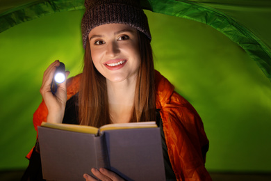 Photo of Young woman with flashlight reading book in tent