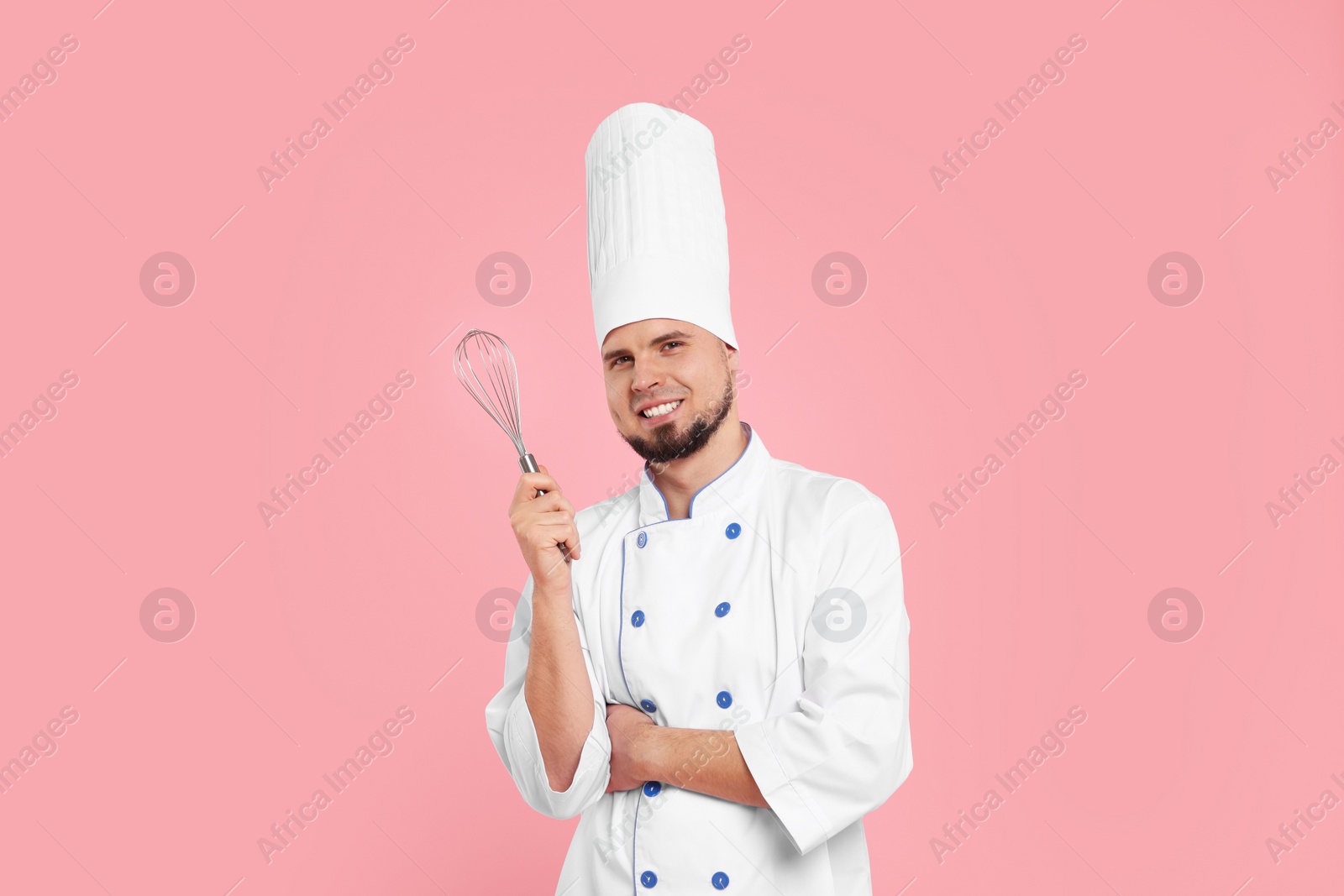 Photo of Happy professional confectioner in uniform holding whisk on pink background