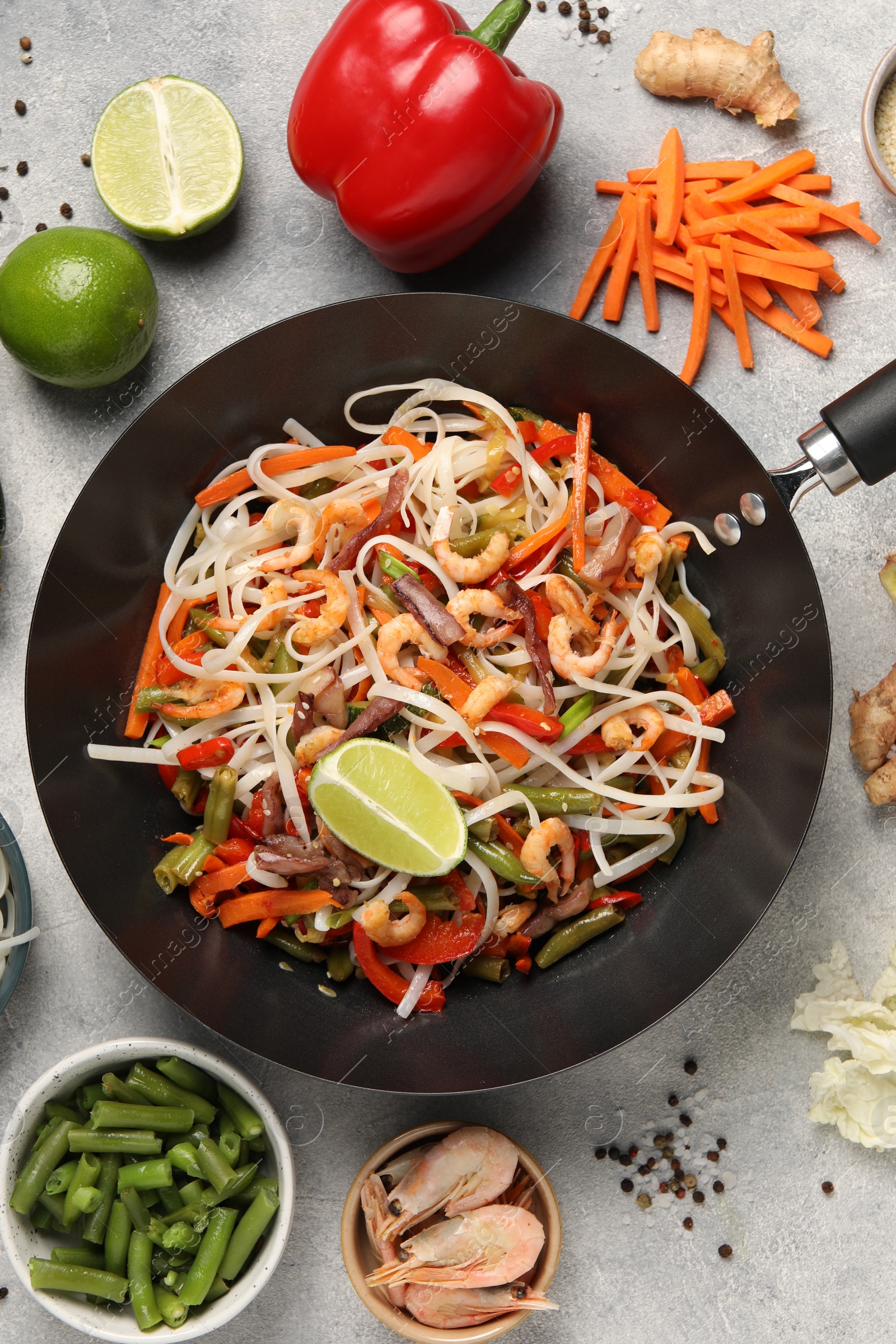 Photo of Shrimp stir fry with noodles and vegetables in wok surrounded by ingredients on grey table, flat lay