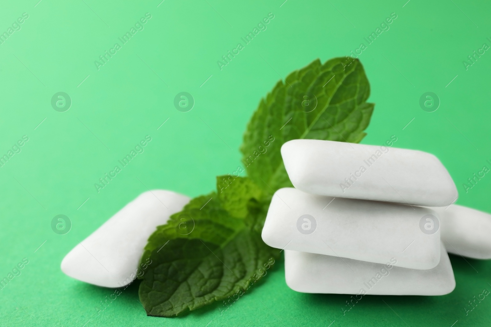 Photo of Tasty white chewing gums and mint leaves on green background, closeup
