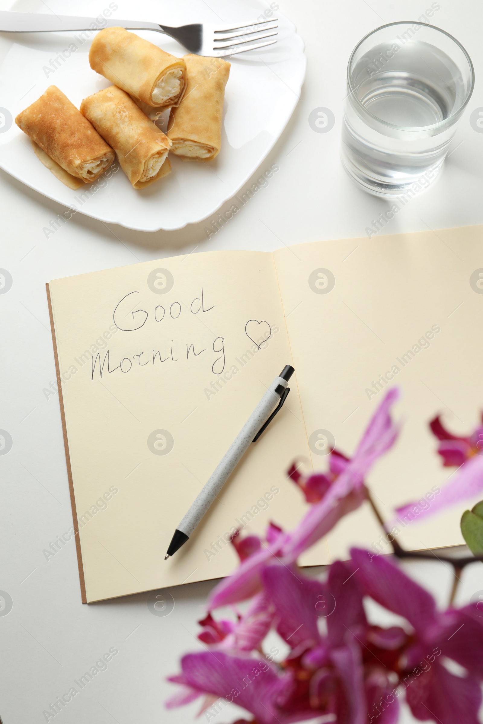 Photo of Notebook with inscription Good Morning, stuffed crepes and glass of water near beautiful blooming orchid on white table, above view