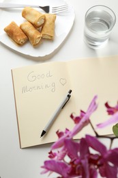 Photo of Notebook with inscription Good Morning, stuffed crepes and glass of water near beautiful blooming orchid on white table, above view