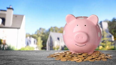 Image of Piggy bank and coins on stone surface and blurred view of beautiful houses, space for text. Mortgage concept