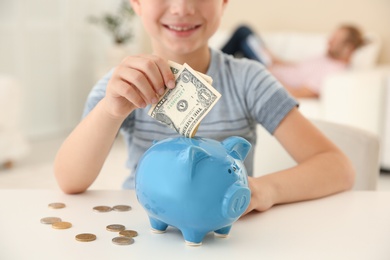 Little boy putting money into piggy bank on table indoors