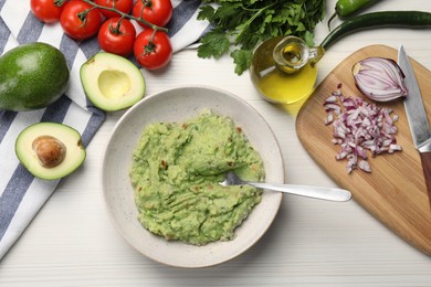 Delicious guacamole in bowl and ingredients on white wooden table, flat lay