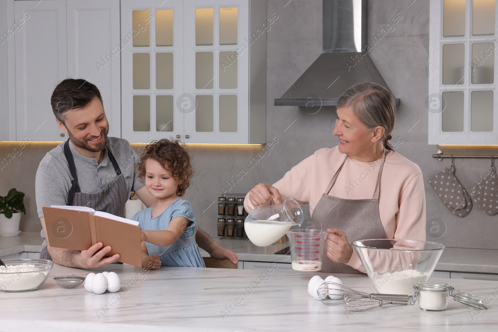 Photo of Cute little girl with her father and grandmother cooking by recipe book in kitchen