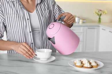 Woman pouring hot water from modern electric kettle in cup indoors, closeup