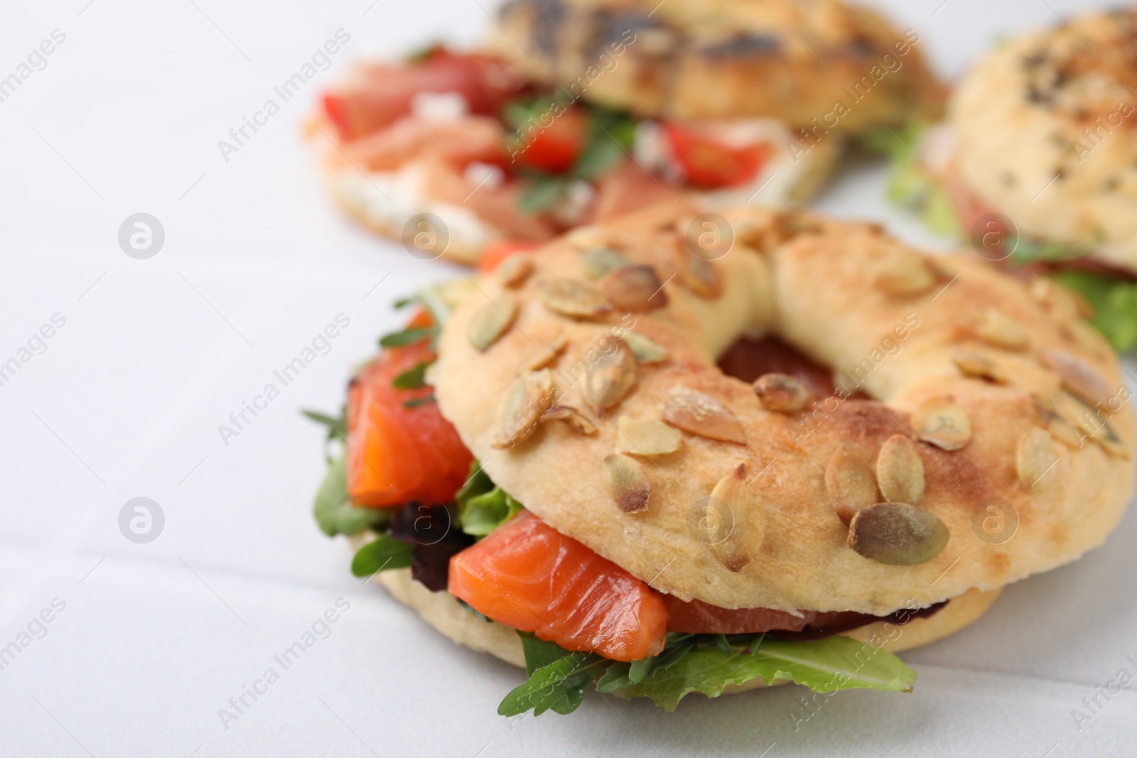 Photo of Tasty bagel with salmon and salad mix on white tiled table, closeup