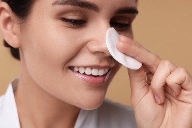 Young woman cleaning her face with cotton pad on beige background, closeup