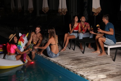 Happy young friends with refreshing cocktails relaxing near outdoor swimming pool at night