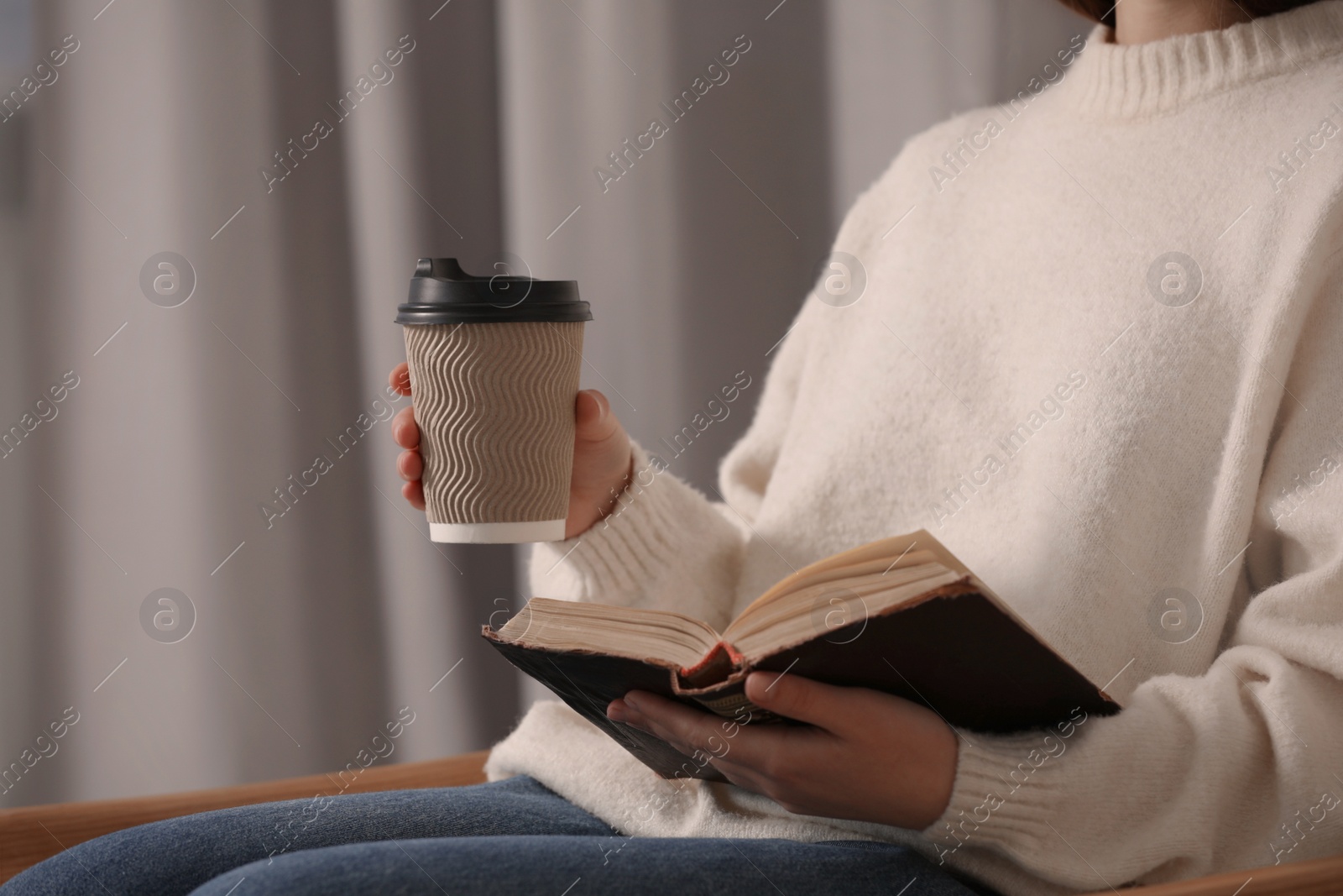 Photo of Woman holding takeaway cardboard cup and book indoors, closeup. Coffee to go