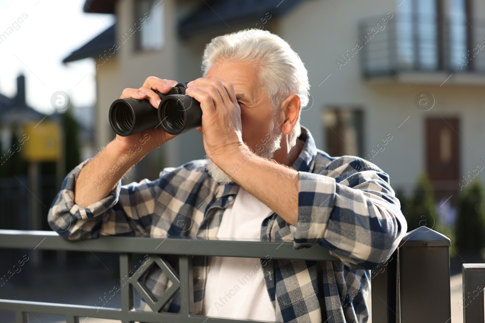 Photo of Concept of private life. Curious senior man with binoculars spying on neighbours over fence outdoors