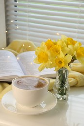 Beautiful yellow daffodils in vase, book and cup of coffee on windowsill. Space for text