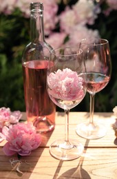 Bottle and glasses of rose wine near beautiful peonies on wooden table in garden