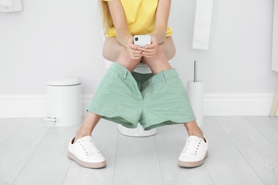 Photo of Young woman using mobile phone while sitting on toilet bowl at home