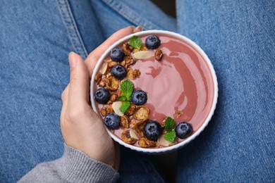Woman holding bowl of delicious fruit smoothie with fresh blueberries and granola, top view