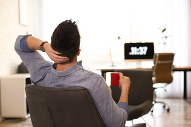 Young man with cup of drink relaxing in comfortable chair at workplace