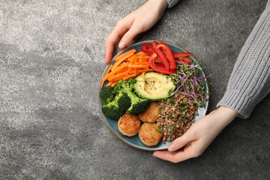 Photo of Woman holding delicious vegan bowl with cutlets, buckwheat and avocado at grey table, top view. Space for text