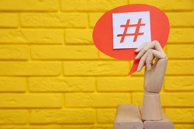 Photo of Wooden mannequin hand holding paper speech bubble with hashtag symbol near yellow brick wall. Space for text
