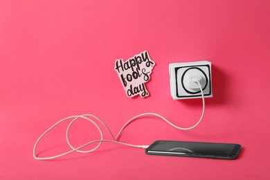 Mobile phone connected to fake plug and words Happy Fool's Day on pink background