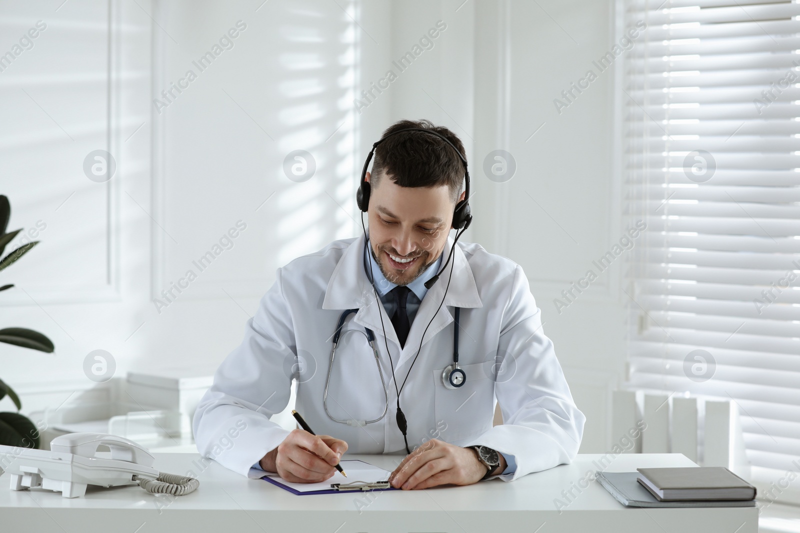 Photo of Doctor with headset consulting patient over phone at desk in clinic. Health service hotline