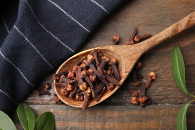 Spoon with aromatic cloves and green leaves on wooden table, flat lay