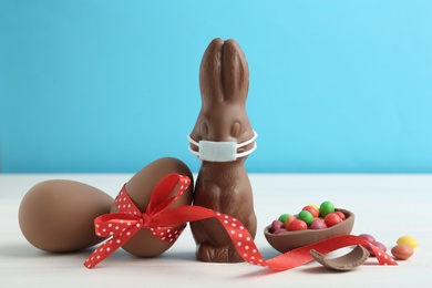 Photo of Chocolate bunny with protective mask and eggs on white wooden table. Easter holiday during COVID-19 quarantine