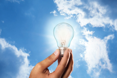 Image of Solar energy concept. Man holding glowing light bulb against blue sky with clouds, closeup