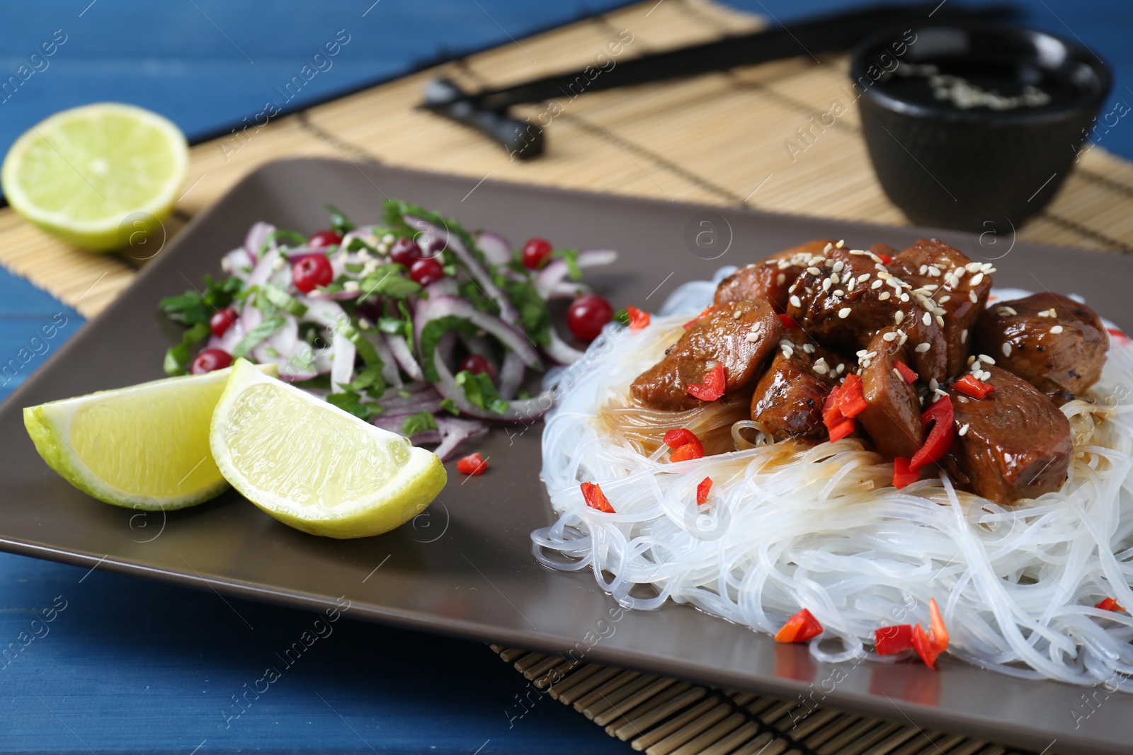 Photo of Pieces of soy sauce chicken with noodle, salad and lime served on blue table, closeup