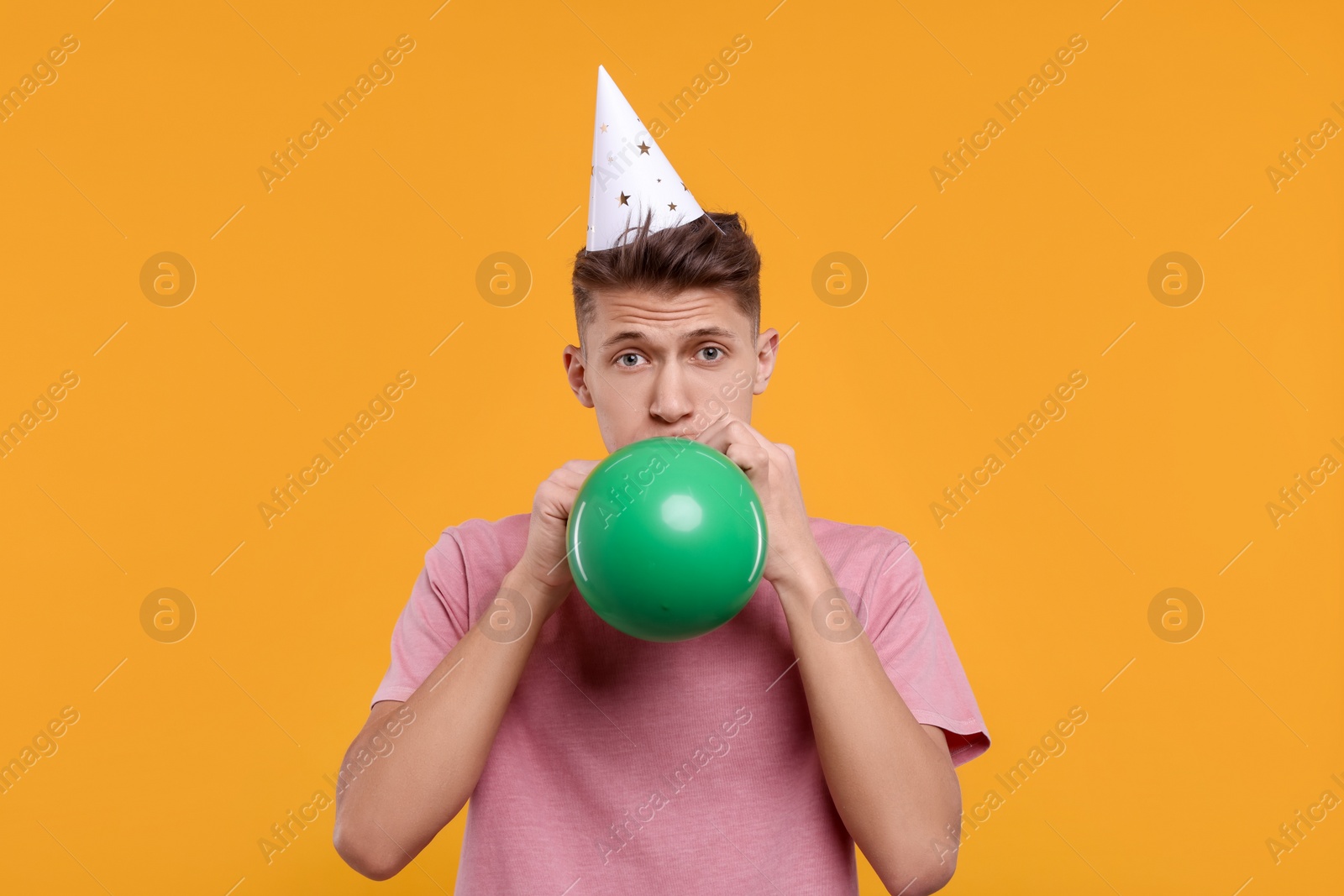 Photo of Young man in party hat inflating balloon on orange background