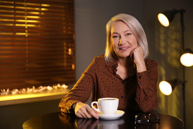 Photo of Beautiful mature woman with cup of coffee indoors. Space for text