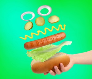 Woman making hot dog on green gradient background, closeup. Ingredients levitating over bun