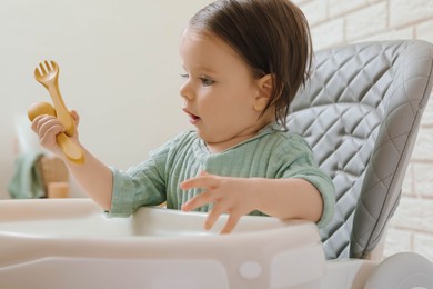 Photo of Cute little baby with cutlery sitting in high chair indoors