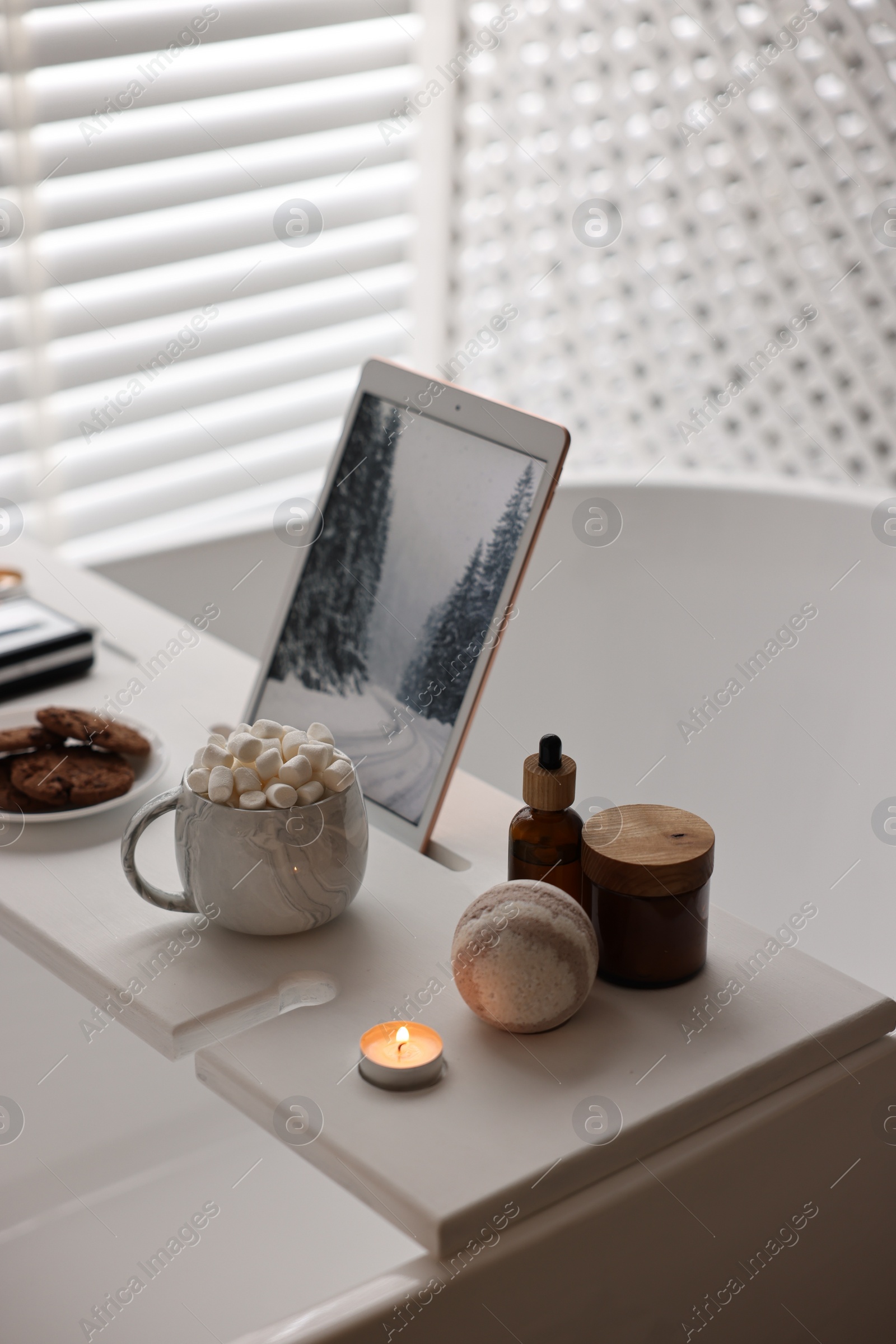 Photo of White wooden tray with tablet, spa products, hot drink and burning candle on bathtub in bathroom