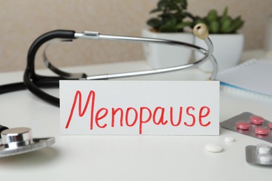 Photo of Card with word Menopause, pills and stethoscope on white table