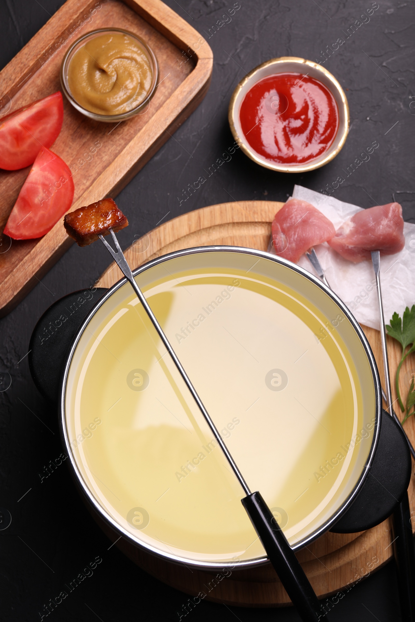 Photo of Fondue pot, forks with meat pieces and other products on black textured table, flat lay