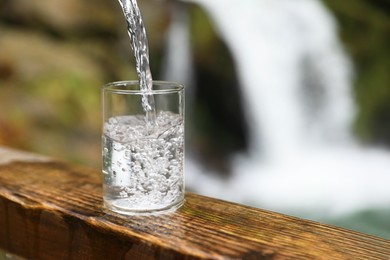 Photo of Fresh water pouring into glass on wooden surface near waterfall outdoors, closeup. Space for text