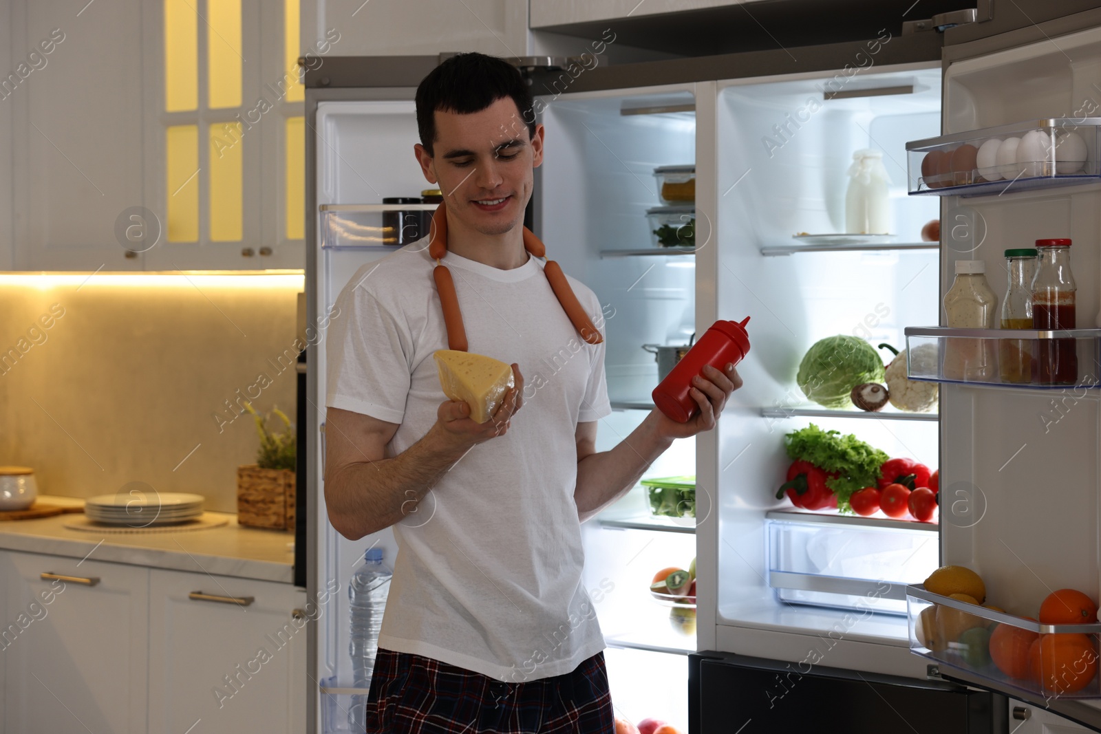 Photo of Happy man with ketchup, cheese and sausages near refrigerator in kitchen