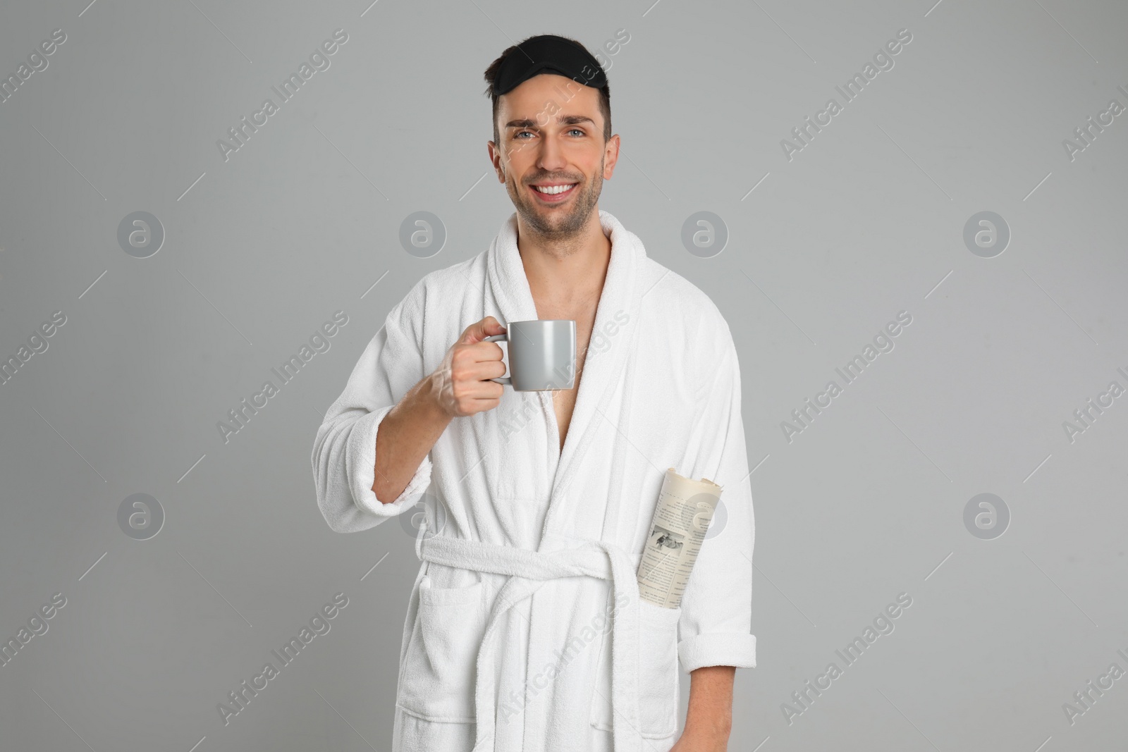 Photo of Happy young man in bathrobe with cup of coffee and newspaper on light grey background