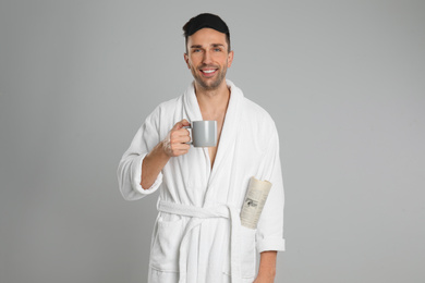 Photo of Happy young man in bathrobe with cup of coffee and newspaper on light grey background