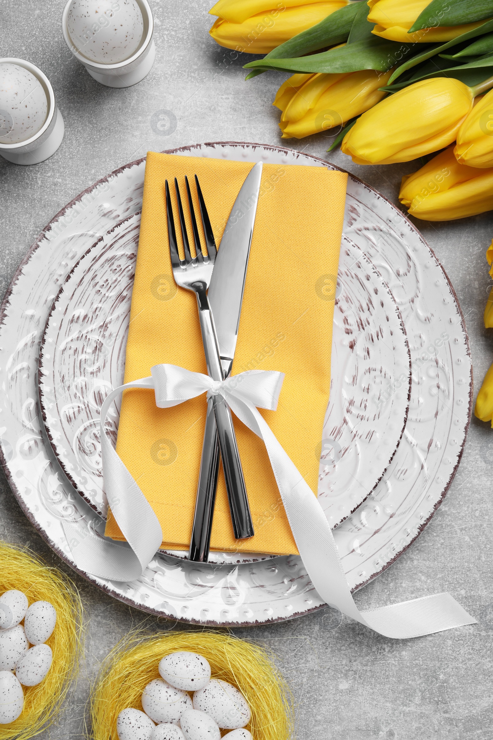 Photo of Festive table setting with tulips and painted eggs on light grey background, flat lay. Easter celebration