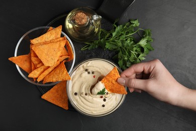 Photo of Woman dipping nacho chip into hummus at black table, top view