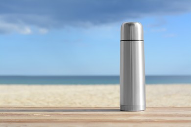 Photo of Metallic thermos with hot drink on wooden surface near sea, space for text