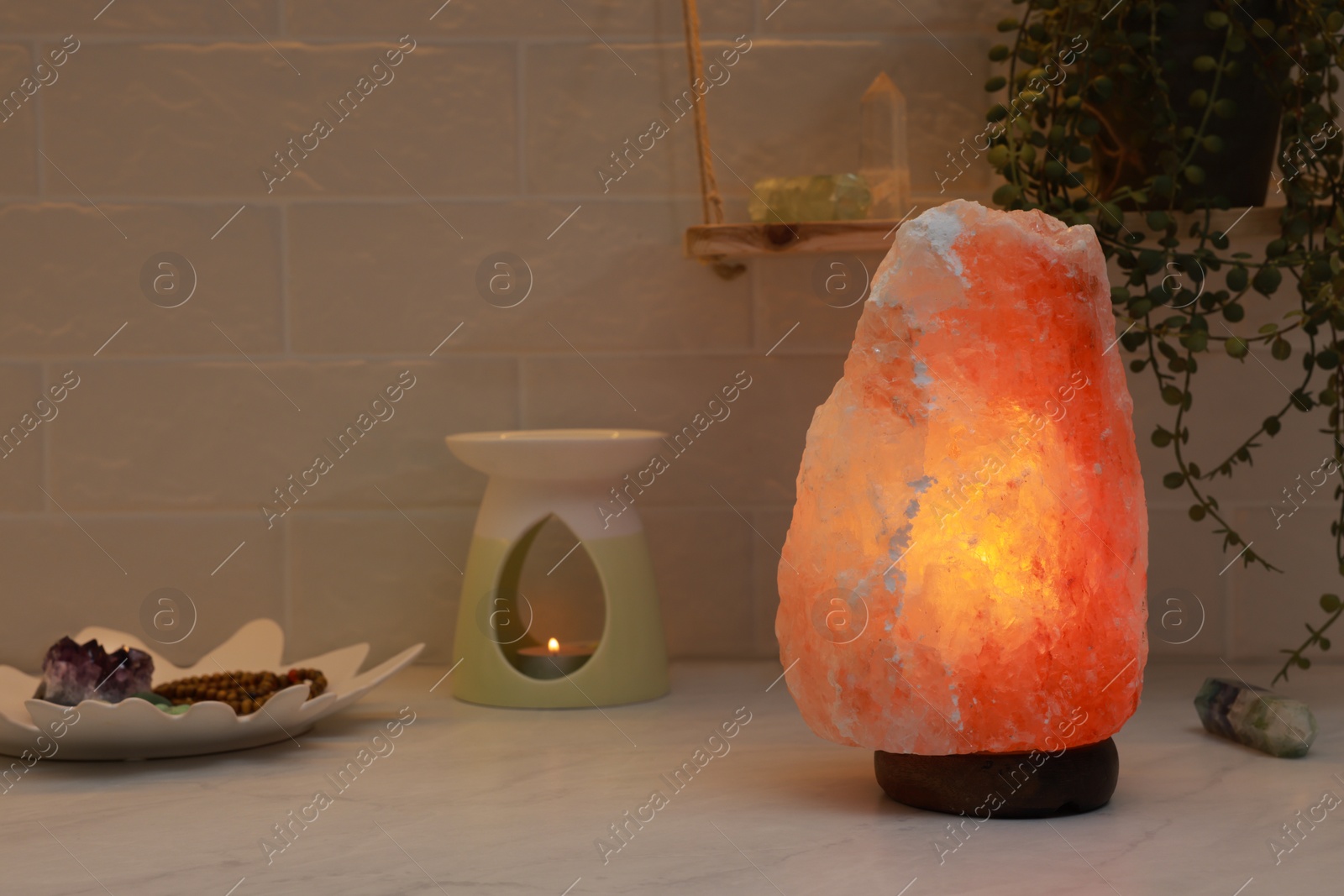 Photo of Himalayan salt lamp, crystals and oil diffuser on white table near brick wall