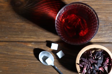 Delicious hibiscus tea in glass, sugar cubes and dry roselle petals on wooden table, flat lay. Space for text