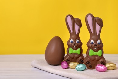 Photo of Chocolate Easter bunnies and eggs on white wooden table against yellow background. Space for text